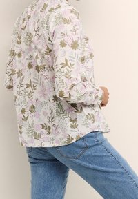 Hvid blomstret bluse med pink og olivengrønne mønstre, lange ærmer, knapmanchetter og en buet kant, parret med lyseblå jeans.