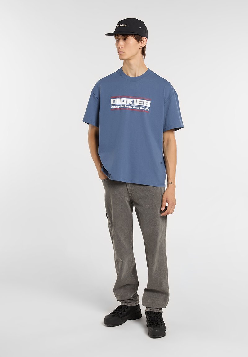 Blue cotton t-shirt with white and red Dickies logo, paired with gray pants and black sneakers; model wearing a black cap.