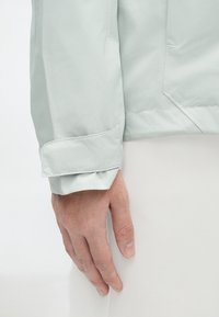 Casaco impermeável verde-claro com uma textura suave, punhos dobrados e um design minimalista. Os detalhes incluem uma costura subtil e um bolso.