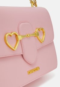 Borsa a tracolla in ecopelle rosa con hardware a forma di cuore dorato e chiusura, con una texture liscia e un logo prominente nella parte inferiore.