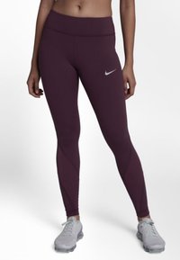 Kobieta w bordo legginsach Nike i jasnoszarych sneakersach, stojąca z nogami rozstawionymi na białym tle.