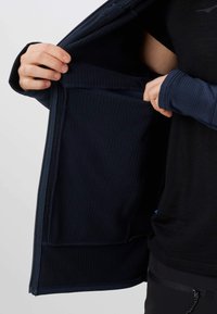 Mains tenant une veste zippée bleu marine ouverte, montrant une grande poche intérieure et une doublure en tissu côtelé.