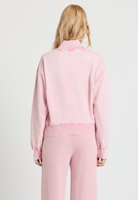 Blonde Person von hinten, trägt ein pastellrosa Sweatshirt und passende lockere Hosen vor einem einfarbigen weißen Hintergrund.