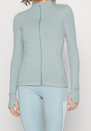 Vrouw draagt een lichtgroene sportjas met ritssluiting en duimgaten, gecombineerd met bijpassende legging met drie witte strepen aan de zijkant.