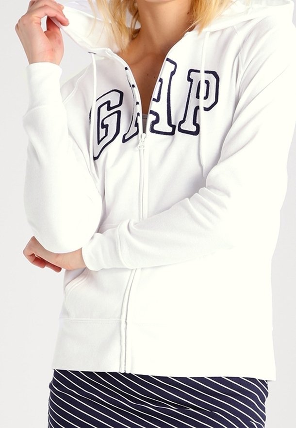 Mujer vestida con una sudadera blanca de GAP con cremallera y letras en azul marino, sosteniendo la capucha con una mano, falda a rayas en azul marino y blanco visible por debajo.