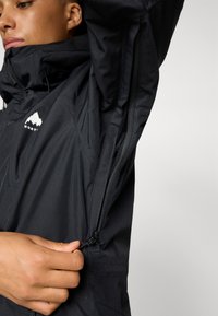 Jachetă neagră cu guler înalt, fermoar parțial și logo de munte. Materialul pare neted și ușor, cu un design elegant și ajustat.