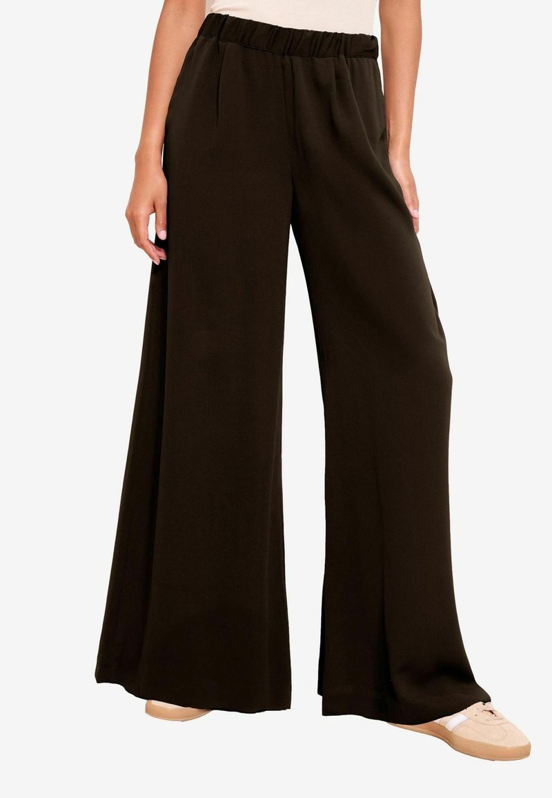 Modèle portant un pantalon large noir ample avec une ceinture élastique et des baskets beiges, debout devant un fond uni.