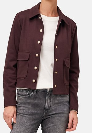 Person trägt eine kurze, dunkelviolette Jacke mit Knöpfen und Klappentaschen über einem weißen Hemd und ausgewaschenen grauen Jeans.