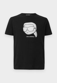 Čierne bavlnené tričko s bielou grafikou stylizovanej postavy v veľkých slnečných okuliaroch, s textom "KARL LAGERFELD" pod obrázkom.