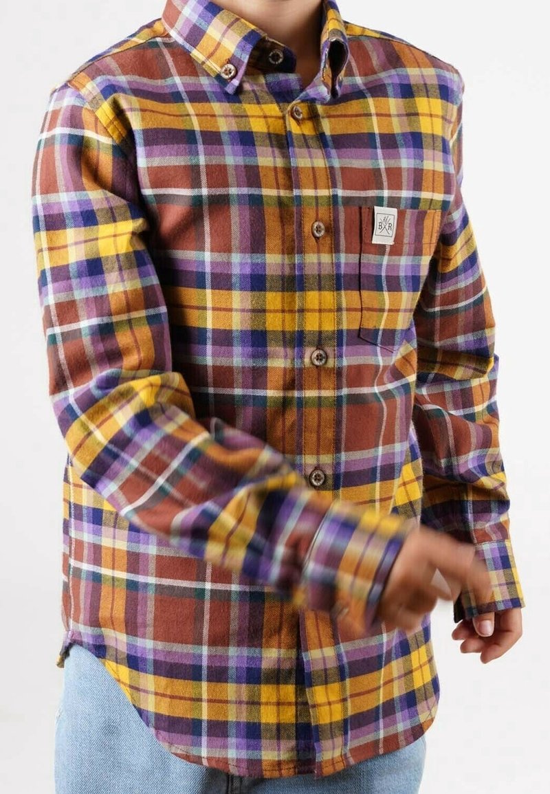 Enfant portant une chemise à carreaux jaune, marron, violet et bleu avec une poche à l'avant et un jean bleu, en pleine action avec des mains floues.