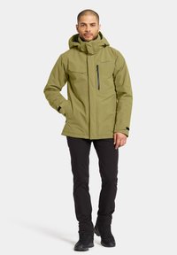 Veste imperméable vert olive avec capuche, poches zippées et coupe décontractée. Assortie avec un pantalon noir et des bottes de randonnée noires.