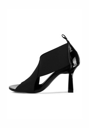 Zapato de tacón alto negro con punta abierta, con tacón de charol y parte superior de textil elástico que presenta un diseño entrecruzado. Un acento en bucle en la parte trasera.