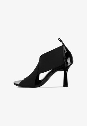 Crne otvorene štikle s visokom petom, od lakirane kože, s gornjim dijelom od elastičnog tekstila koji ima dizajn u obliku križa. Ima naglašeni detalj u obliku petlje straga.