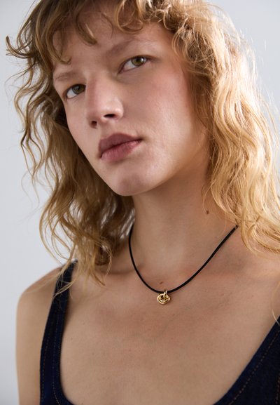 Collier en cordon noir ornée d'un pendentif circulaire en or, porté contre un décolleté nu sur un fond en tissu bleu foncé.
