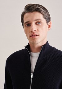 Pull en maille côtelée bleu marine avec un col montant et une fermeture éclair. Porté sur un t-shirt de couleur claire, présentant une texture lisse.