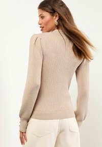 Beige gerippter Pullover mit langen Ärmeln, taillierter Passform und dezenten Puffschultern, mit Knopfdetails an den Ärmeln. Weiche Textur.