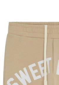 Pantalons de survêtement beige en tissu doux, avec un texte graphique blanc "SWEET" en lettres majuscules, et une taille élastique avec cordon.