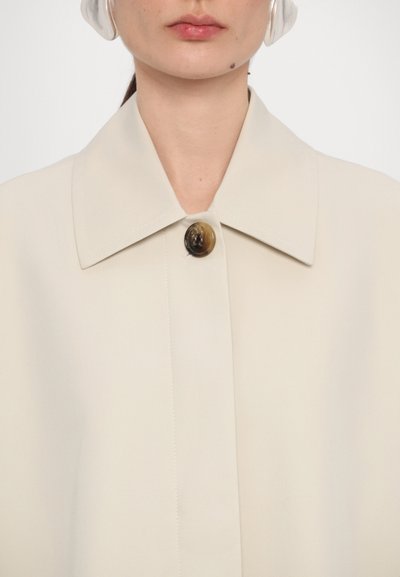 Light beige outerwear with a wide collar, featuring a large, round brown button at the neckline; smooth, crisp fabric texture.