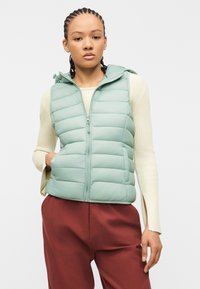 Νέος με πλεξούδες, ντυμένος με ανοιχτό πράσινο γιλέκο γεμισμένο επάνω σε ένα κρεμ πουλόβερ και καφέ-κόκκινο παντελόνι, στέκεται με το ένα χέρι στην τσέπη.