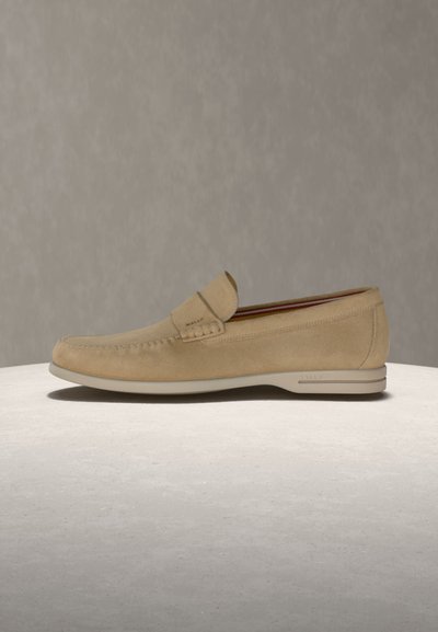 Μπεζ ανδρική μοκασίνια από σουέντ με ραφές τύπου moc toe, χαμηλή ελαστική σόλα και λεπτομέρεια με λουρί στο πάνω μέρος, εμφανιζόμενα σε φωτεινή επιφάνεια.