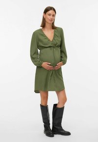 Grūtniece zaļā garā piedurknē tērptā kleitā tur vēderu, vilkusi melnas augstus zābakus, stāv uz vienkārša balta fona.