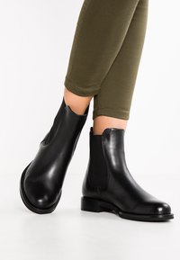 Black leather ankle boots with elastic side panels, rounded toe, low heel, and smooth texture. Worn with olive green fitted pants.