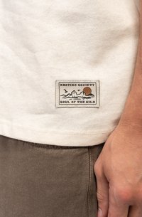 T-shirt en coton beige avec une étiquette en tissu rectangulaire portant le texte "Knotiko Society" et "Soul of the Wild", ainsi que des éléments de design graphique.