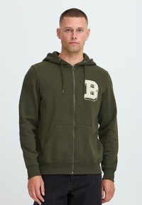 Muž oblečený v olivovo zelenej bundě na zips s velkým bielym "B" na ľavej hrudi a čiernych nohaviciach, stojaci pred bielym pozadím.
