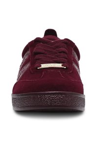 Burgundy suede sneakers with flat laces, padded tongue, side stripe detail, and textured rubber sole. Gold hardware accent on tongue.