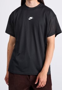 Persona vestida con una camiseta de malla negra de Nike con el logo centrado en el pecho y pantalones de pana marrones, de pie contra un fondo liso.