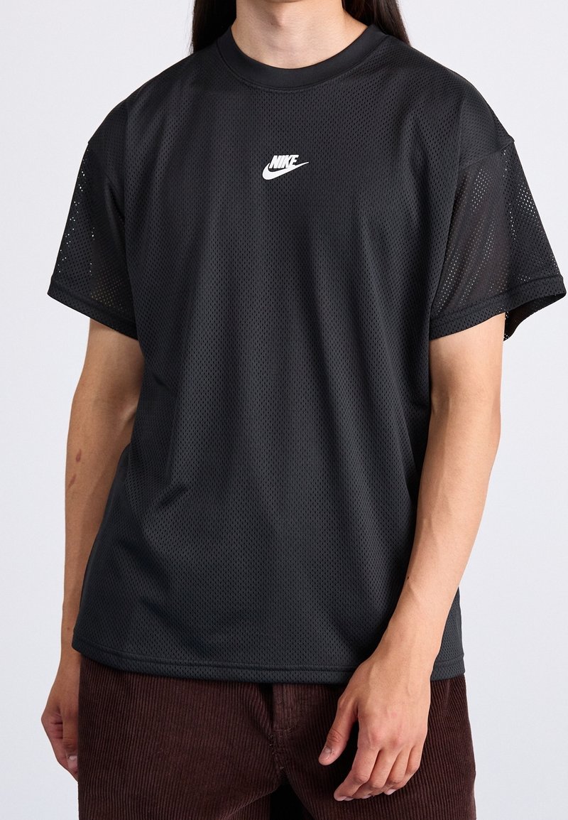 Persona vestida con una camiseta de malla negra de Nike con el logo centrado en el pecho y pantalones de pana marrones, de pie contra un fondo liso.