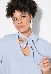 Blusa azzurro chiaro con collo annodato, caratterizzata da scollatura a V e una texture di tessuto liscia. Orecchini bianchi ovali e grandi completano il look.