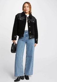 Giacca in shearling nero e pelle con chiusura a bottoni, maniche lunghe e colletto, abbinata a jeans a vita alta blu a gamba larga.