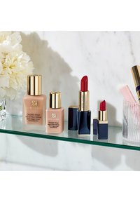 Zwei beige Estee Lauder Double Wear Foundationflaschen mit goldenen Akzenten, neben zwei roten Lippenstiften in blauem und goldenem Verpackungsdesign, auf einem Glastablett.