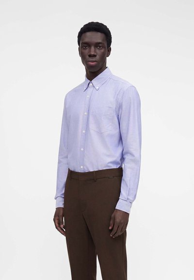 Homme adulte portant une chemise bleu clair à manches longues boutonnée et un pantalon marron foncé, debout devant un fond blanc uni.