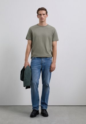 Man in een olijfgroen t-shirt en blauwe spijkerbroek, met een donkergroene jas in zijn linkerhand, staand tegen een effen lichte muur.