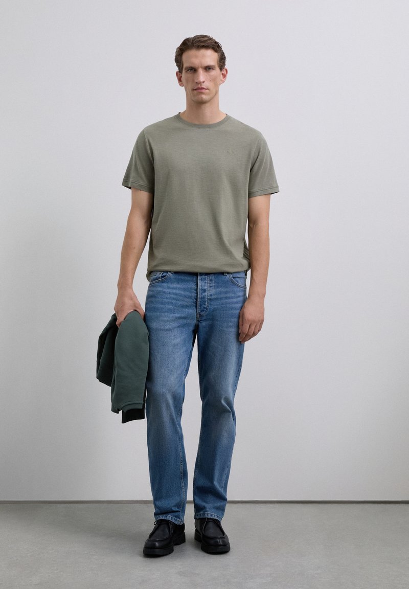 Man wearing olive-green t-shirt and blue jeans, holding a dark green jacket in left hand, standing against a plain light wall.