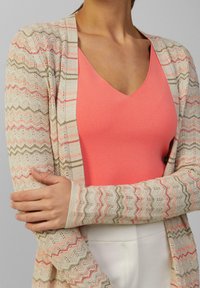 Leichte, strukturierte Strickjacke in Beige mit rosa und grünen Zickzackmustern, über einem korallenfarbenen V-Ausschnitt-Oberteil getragen.