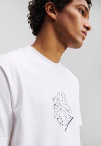 Balta medvilnės marška su juodu siuvinėjimu ant krūtinės, primenančiu ranką, su tekstu "KARL LAGERFELD JEANS" žemiau.
