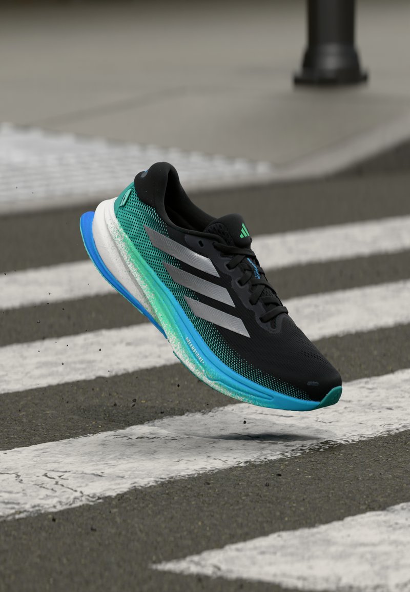 Chaussure de course Adidas noire et bleu sarcelle en lévitation au-dessus d'un passage piéton sur une rue pavée, montrant le mouvement et les détails de la semelle usée.