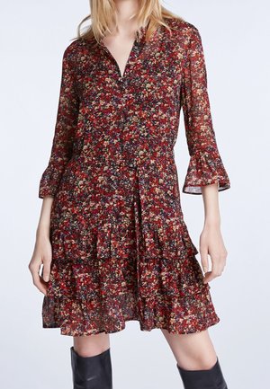 Frau trägt ein langärmeliges, rotes Blumenkleid mit Stufen und Rüschen, kombiniert mit schwarzen kniehohen Stiefeln, stehend vor einem einfarbigen Hintergrund.