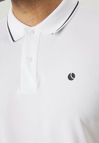 Weißes Poloshirt aus strukturiertem Stoff, mit einem schwarz gestreiften Kragen und einem kleinen schwarzen Logo auf der linken Brust.