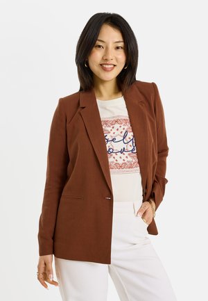 Jeune femme souriante, portant un blazer marron sur une chemise blanche imprimée, un pantalon blanc et des boucles d’oreilles créoles dorées, debout devant un fond uni.