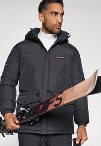Homem com um casaco de inverno Berghaus preto, segurando um único esqui rosa e preto com fixações Tyrolia, olhando para o lado contra um fundo simples.