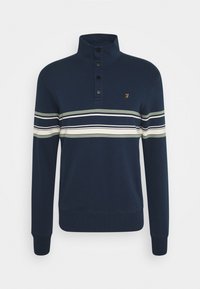 Tmavě modrý svetr s vysokým límcem a čtyřmi černými knoflíky, vodorovné bílé a zelené pruhy na hrudi a horních pažích, malé žluté logo.