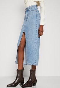 Gonna di jeans azzurro chiaro con spacco frontale, vita alta e taglio dritto. Abbinata a stivali alla caviglia marrone scuro con punta squadrata.