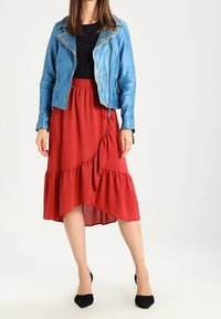 Frau trägt eine blaue Lederjacke, ein schwarzes Oberteil, einen roten, gerüschten Midirock und schwarze, spitze Absatzschuhe und steht vor einem einfarbigen Hintergrund.