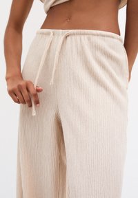 Beżowe, teksturowane spodnie typu wide-leg z elastycznym paskiem w talii, z widocznymi pionowymi paskami na materiale. Ręka umieszczona w pobliżu pasa.