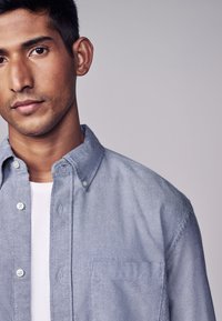 Helles blaues Hemd mit Kragen, weicher Textur und einer Brusttasche, getragen über einem weißen T-Shirt. Schlichtes Design, lässiges Aussehen.