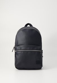 Mochila de cuero negro con un acabado suave, parte superior redondeada, bolsillo frontal con cremallera plateada y un parche de logo en la parte inferior.